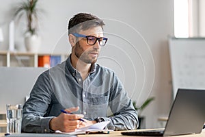 Successful middle aged businessman working at office, sitting at workdesk in front of laptop and taking notes