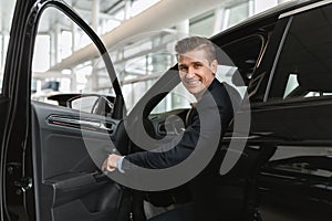 Successful millennial car salesman sitting inside auto salon, looking at camera through open door in modern dealership