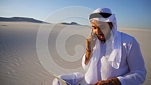 Successful Muslim Arabian UAE Sheikh architect communicates on phone with client sitting on sand with laptop in large