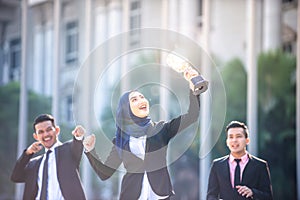 Successful Muslim business woman with arms up holding a trophy and celebrating, with male teammates supporting from behind. Female