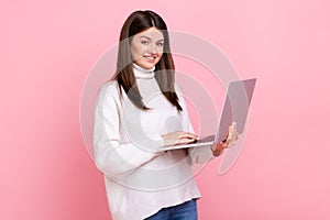 Successful office worker girl looking at camera with happy face, holding laptop, typing on keyboard.