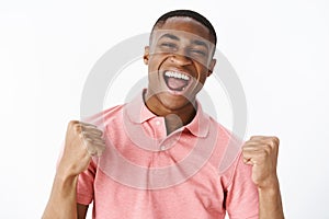 Successful pleased and happy handsome young african american guy in pink shirt clenching fists in victory and cheer