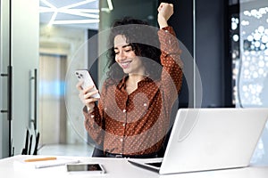 Successful satisfied business woman celebrating success and triumph, boss at workplace holding phone and hand up victory