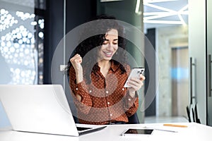 Successful satisfied business woman celebrating success and triumph, boss at workplace holding phone and hand up victory