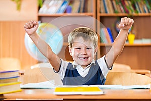 Successful schoolboy with hands up sitting at desk