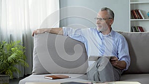 Successful senior man sitting on couch with tablet, using modern gadget for work