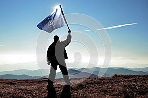 Successful silhouette man winner waving NATO flag on top of the mountain