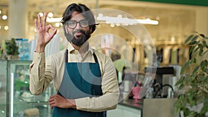 Successful small business owner bearded Indian Arabian man in eyeglasses standing in cafeteria restaurant showing ok