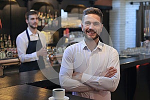 Successful small business owner standing with crossed arms with employee in background preparing coffee