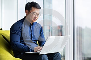 Successful and smiling asian man working with laptop inside office with notebook, senior businessman in glasses and