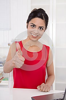 Successful smiling asian secretary with thumb up at desk at office.