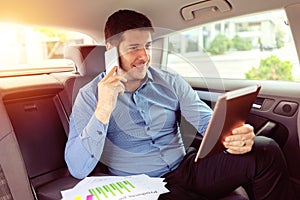 Successful smiling businessman talking on phone while sitting in back seat of car using tablet