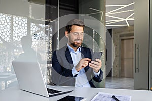 A successful and smiling young male businessman is sitting in the office at a desk in a suit and using a mobile phone