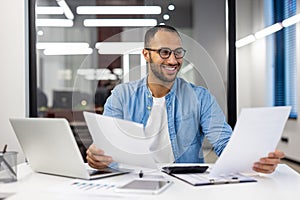 A successful and smiling young Muslim man in glasses is sitting in the office at a desk with a laptop and working with