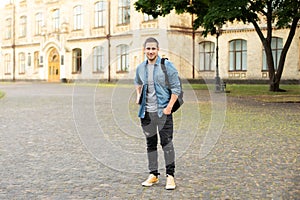 Successful student is standing with backpack and laptop behind park near campus. Smile young man standing against university