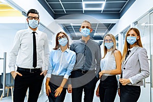 Successful team of business workers, wearing medical protective masks on their face, posing together in a modern office, happy