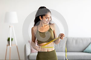 Successful weight loss concept. Fit young Indian woman in sports clothes measuring waist with tape at home