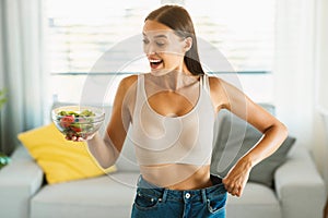 Successful weight loss diet. Excited slim lady holding bowl with vegetable salad and showing result of slimming