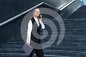 Successful woman talking on mobile phone, holding laptop on background steps