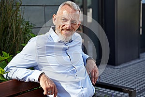 Successful worker. Portrait of confident, cheerful middle aged businessman smiling aside while sitting on the bench