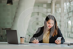 Successful young Asian businesswoman holding a smartphone working on a laptop computer financial graph data on the desk in the off