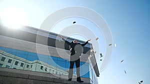 Successful young business man boss winner stands outdoors near blue glass building, wears stylish black suit, throws