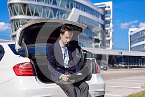 Successful young businessman freelancer working outside in the city. man sit on car boot with tablet