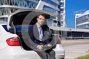 Successful young businessman freelancer working outside in the city. man sit on car boot with tablet