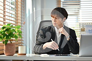 Successful young businessman sitting at workplace and looking through window, pondering idea or developing business