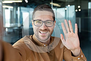 Successful young businessman talking with friends on video call, man smiling and waving at smartphone camera, worker at