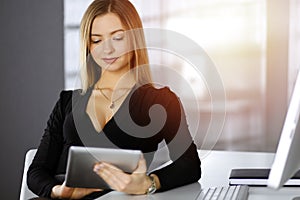 Successful young businesswoman is using a computer tablet, while working in a sunny cabinet of her firm. Business