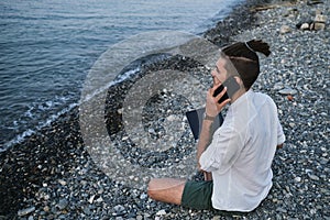 Successful young Caucasian male freelancer is sitting on beach and remotely working online using laptop. Businessman calls on the