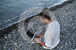 Successful young Caucasian male freelancer is sitting on beach and remotely working online using laptop. Hipster businessman works