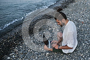 Successful young Caucasian male freelancer is sitting on beach and remotely working online using laptop. Hipster businessman works