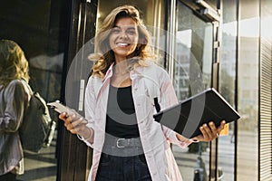 Successful young caucasian woman smiles teeth at camera, holds technological devices on street.