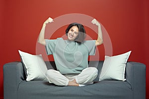 Successful young girl showing power raised arms, demonstrate biceps muscles proud of achievements, sitting on sofa