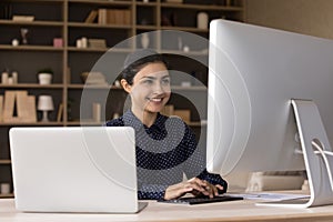Successful young Indian female work at desk using two computers