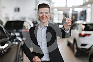 Successful young salesman showing car key at camera in auto dealership, selective focus. Vehicle local distribution