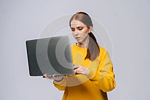 Successful young woman student holding laptop computer and typing on isolated gray background.