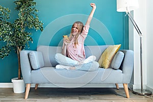 Successful young woman using her mobile phone and celebrating something while sitting on sofa at home