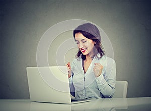 Successful young woman winner with arms raised looking at laptop sitting at desk