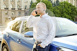 Successfull happy young man and his car in soft sunset light on urbanistic background. Busines man with vehicle on roadside.