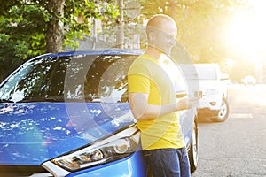 Successfull happy young man and his car in soft sunset light on urbanistic background. Busines man with vehicle on roadside.