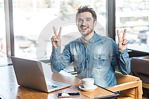 Successfull positive bearded young freelancer in blue jeans shirt are sitting in cafe and working on laptop with toothy smile and