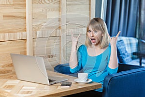 Successfull young girl freelancer with blonde hair in blue blouse are sitting in cafe and working on laptop with toothy smile and