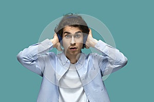 Suddenness. Portrait of a young beautiful man wearing white t-shirt and blue shirt in headphones covers ears with hands opened