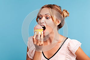 Sugar addiction. Portrait of hungry blonde young adult woman in dress biting delicious cake, looking with desire to eat sweet