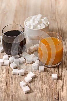 Sugar cubes are scattered on the kitchen counter. There is cola in one glass and juice in the other. In the background a sugar bow