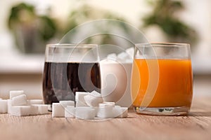 Sugar cubes are scattered on the kitchen counter. There is cola in one glass and juice in the other. In the background a sugar bow
