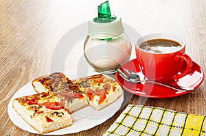 Sugar, pieces of khachapuri with tomato and basil in white plate, coffee in cup, spoon on saucer, napkin on wooden table
